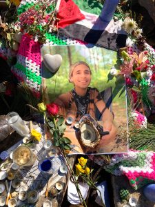 A photo of Tortuguita, a 26-year-old Indigenous, non-binary Venezuelan, on an altar surrounded by candles, shells, and flowers. The photo rests on crocheted white, green, and pink blanket.