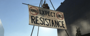 A person and sign in the air, “Expect resistance.” Two red crossed out circles either side of the message, one circle saying “Enbridge” and the other “Line 3.”