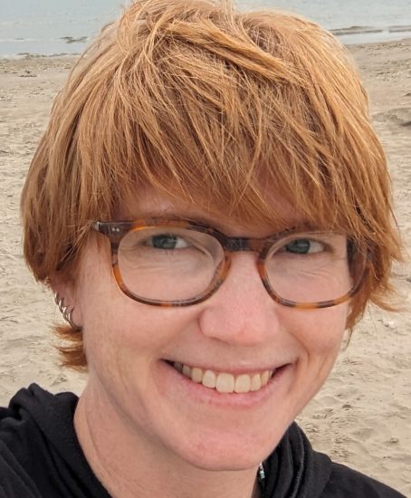 Heather smiling with sand and water behind her. She has pale skin, short red hair with bangs that touch her tortoiseshell glasses, wearing a black top