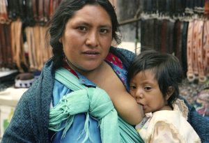 A women is looking at the camera, with a child suckling at her breast.
