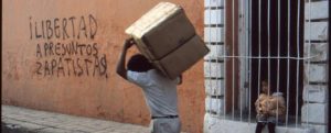 A person bent over, carrying a large cardboard box on their shoulders, as seen from behind. Spray painted on a stucco wall in front of them reads libertad a presuntos zapatistas, meaning freedom for alleged zapatistas